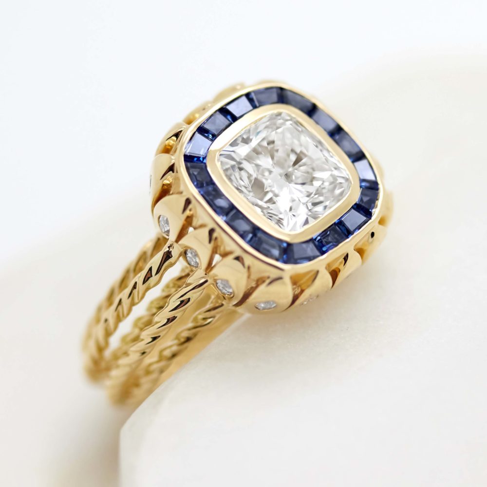 Radiant cut diamond with a blue sapphire bezel set halo set upon a rope gold band